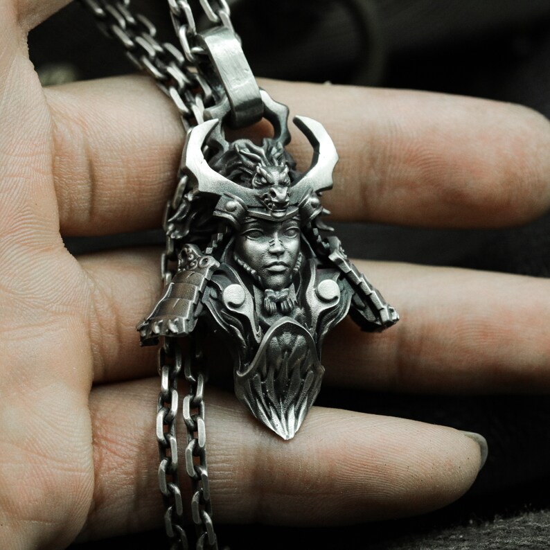 Puede incluir: Un collar con un colgante de plata que presenta una escultura detallada de un guerrero samur&aacute;i que lleva un casco con un dise&ntilde;o de drag&oacute;n. El colgante est&aacute; unido a una cadena de plata.