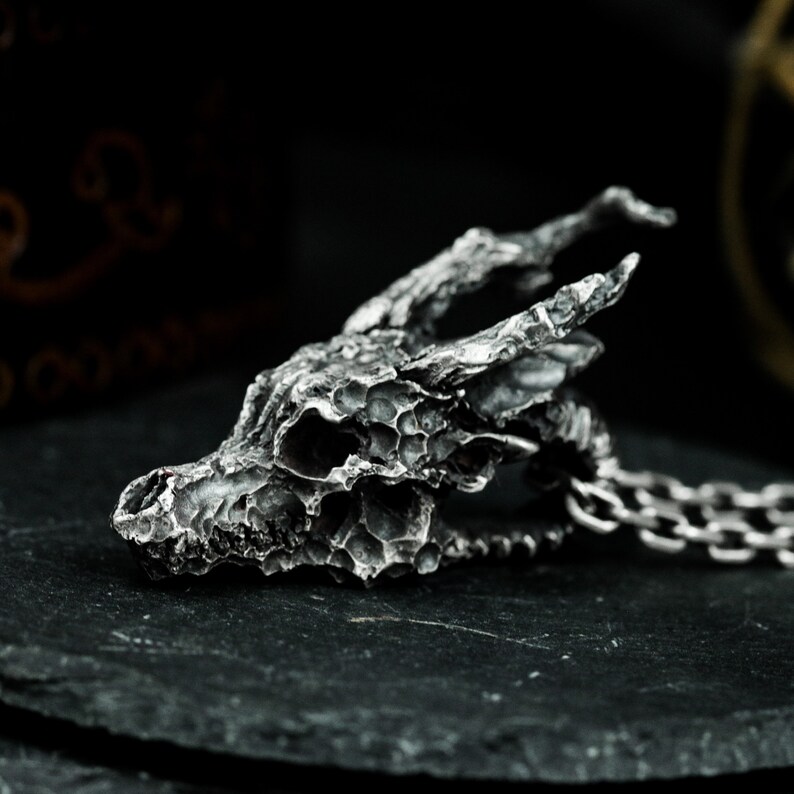 May include: A silver dragon skull pendant with a detailed, textured surface. The pendant is attached to a silver chain.