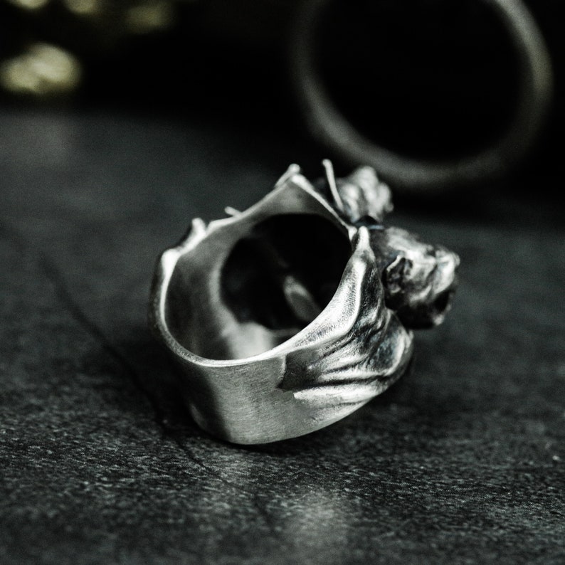 Puede incluir: Un anillo de metal plateado con un dise&ntilde;o de murci&eacute;lago esculpido. La banda tiene un acabado cepillado y oxidado que resalta la textura detallada de las alas y la cabeza del murci&eacute;lago, que envuelven la parte superior de la estructura circular.