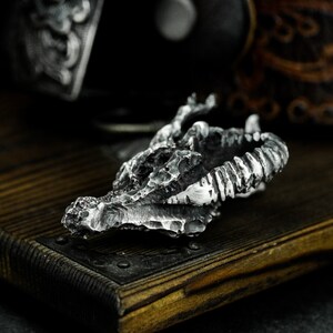 May include: A detailed silver dragon skull pendant with a textured surface. The pendant is made of metal and has a realistic design.