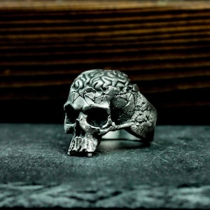 May include: A silver skull ring with a detailed brain design. The skull is cracked and the brain is visible.