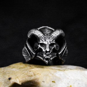 Può includere: Un anello d'argento con un designo di teschio dettagliato con corna. Il teschio ha una superficie testurizzata e sembra essere fatto di metallo.