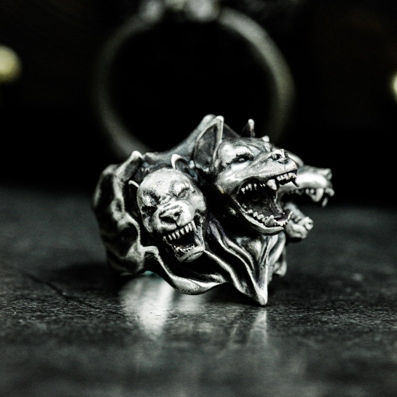 Puede incluir: Un anillo de metal plateado con un dise&ntilde;o detallado de tres cabezas de canino. Cada cabeza muestra dientes descubiertos, orejas puntiagudas y una expresi&oacute;n de gru&ntilde;ido. La superficie met&aacute;lica tiene un acabado oscuro y oxidado definido.