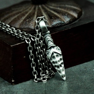 May include: A silver chain necklace with a textured, pointed pendant. The pendant is shaped like a spearhead and has a hammered finish.