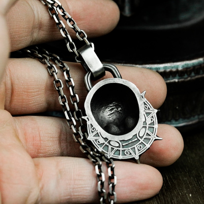 May include: A silver chain necklace with a pendant featuring a detailed, circular design with a raised, textured center. The pendant has a dark, almost black, finish.