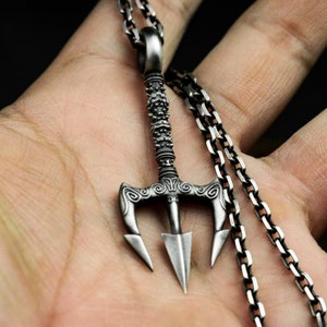 May include: A silver trident pendant necklace with a detailed design. The pendant features a three-pronged spearhead with intricate carvings and a chain link necklace.