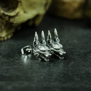 May include: A pair of silver earrings featuring detailed wolf head designs. The earrings are made of metal and have a textured finish.