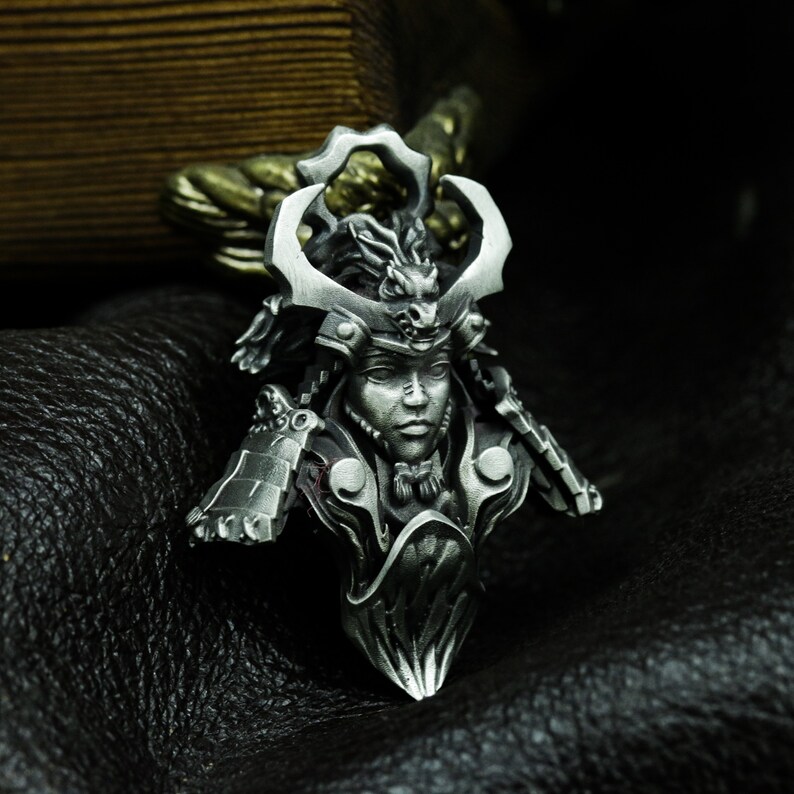 Puede incluir: Un colgante de plata que presenta un busto detallado de un guerrero samur&aacute;i que lleva un casco con un dise&ntilde;o de drag&oacute;n. El colgante est&aacute; elaborado de forma intrincada con una superficie texturizada.