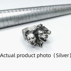 Puede incluir: Un anillo de plata que presenta tres cabezas de perro detalladas con los dientes descubiertos y expresiones de gru&ntilde;ido. El anillo est&aacute; colocado junto a un mandril de metal, que muestra marcas incrementales para medir el tama&ntilde;o en cent&iacute;metros.