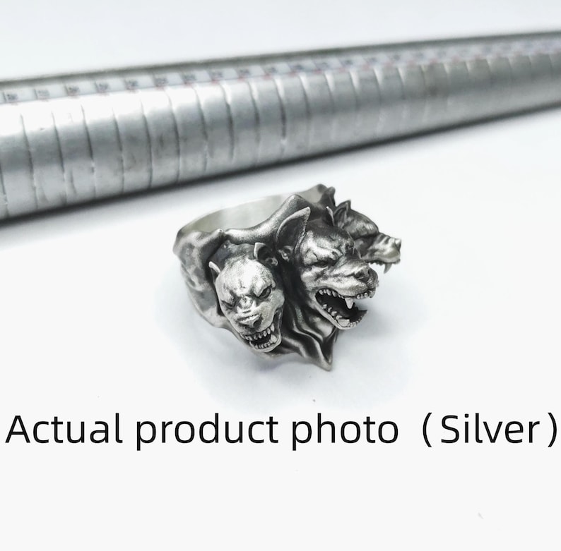 Puede incluir: Un anillo de plata que presenta tres cabezas de perro detalladas con los dientes descubiertos y expresiones de gru&ntilde;ido. El anillo est&aacute; colocado junto a un mandril de metal, que muestra marcas incrementales para medir el tama&ntilde;o en cent&iacute;metros.