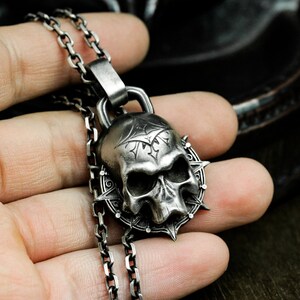 May include: A silver skull pendant necklace with a detailed design and a spiked border. The pendant is hanging from a silver chain.