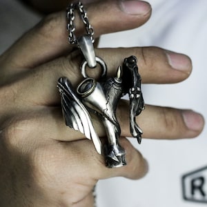 Puede incluir: Un collar con un colgante de plata que presenta un dise&ntilde;o detallado de una criatura alada sosteniendo un objeto estilizado. El colgante tiene una superficie texturizada y un acabado plateado oscuro.