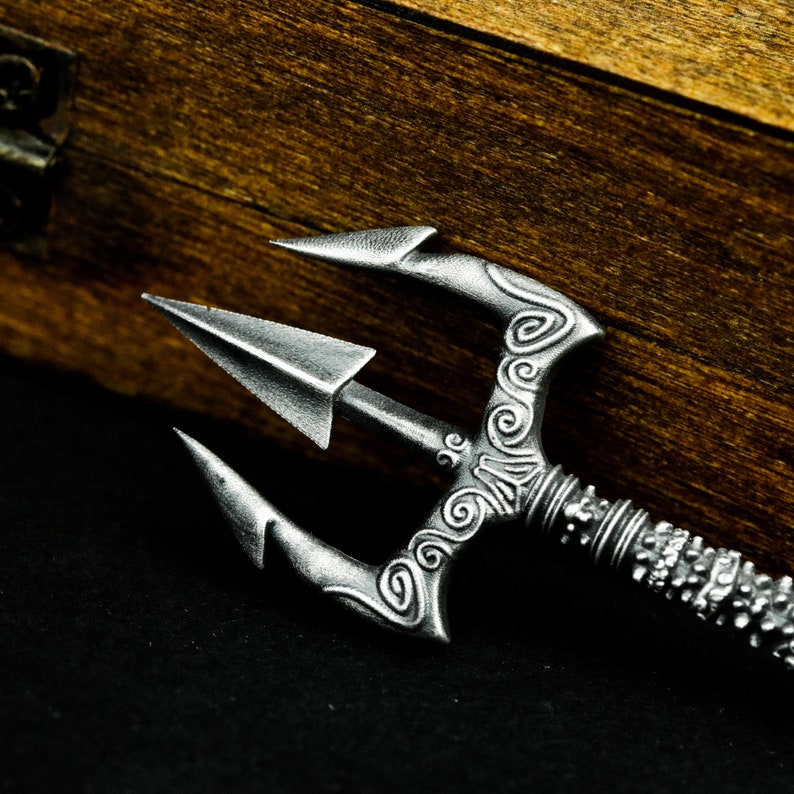 May include: A silver trident with intricate details and a sharp point. The trident is lying on a dark surface.