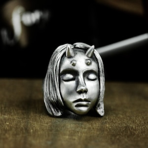 May include: A silver ring with a detailed sculpture of a woman's face with closed eyes and horns. The ring is on a wooden surface.