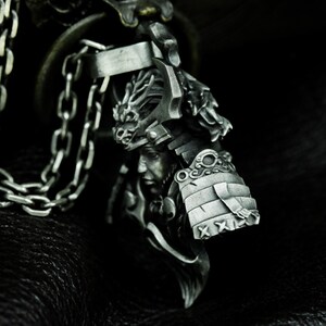 Puede incluir: Un colgante de plata con una escultura detallada de un guerrero samur&aacute;i que lleva un casco con un dise&ntilde;o de drag&oacute;n. El colgante est&aacute; unido a una cadena de plata.