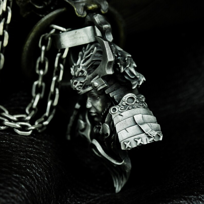Puede incluir: Un colgante de plata con una escultura detallada de un guerrero samur&aacute;i que lleva un casco con un dise&ntilde;o de drag&oacute;n. El colgante est&aacute; unido a una cadena de plata.