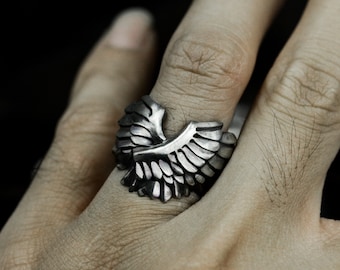 May include: A silver ring with a detailed eagle wing design. The ring is worn on a person's finger.