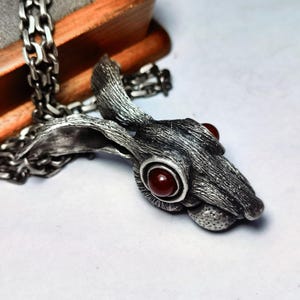 Pode incluir: Um colar com um pingente de prata que representa uma cabeça de coelho detalhada com olhos de pedras preciosas vermelhas. A pelagem do coelho é texturizada e o pingente está preso a uma corrente de prata.