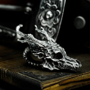 May include: A detailed silver skull ring with a textured, bony design. The skull has a pointed nose and sharp teeth. The ring is set on a dark wooden surface.