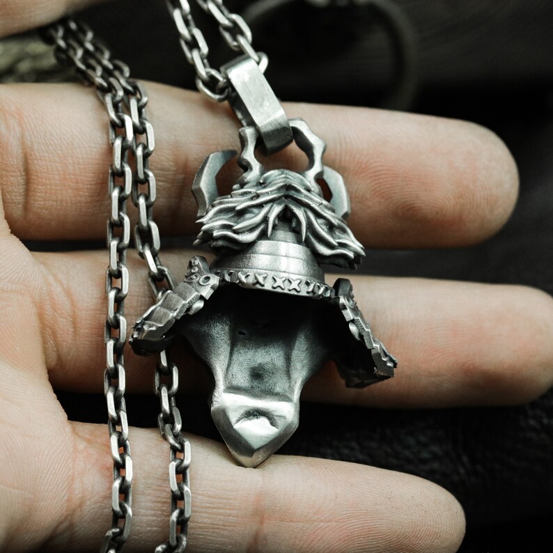 Puede incluir: Un collar de cadena de plata con un colgante detallado de un casco de samur&aacute;i. El casco est&aacute; intrincadamente dise&ntilde;ado con una superficie texturizada y una cresta elevada.
