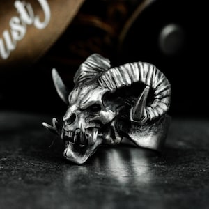 Może przedstawiać: Srebrny pierścionek z detalicznym wzorem czaszki z rogami i ostrymi zębami.