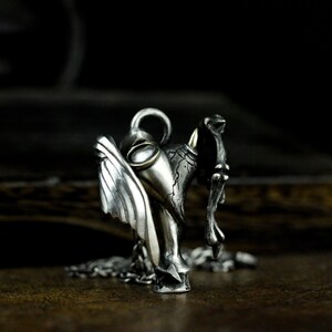 Puede incluir: Un collar con un colgante de plata que presenta una escultura detallada de un &aacute;ngel con alas y un demonio con cuernos. El colgante tiene un peque&ntilde;o acento redondo de color dorado.