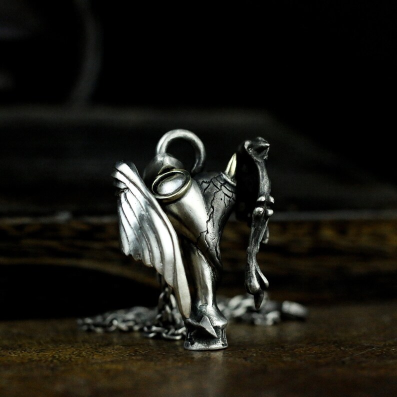 Puede incluir: Un collar con un colgante de plata que presenta una escultura detallada de un &aacute;ngel con alas y un demonio con cuernos. El colgante tiene un peque&ntilde;o acento redondo de color dorado.