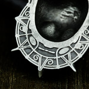 May include: A silver-colored metal pendant with a circular design featuring intricate details and a raised center. The pendant has a sharp, pointed edge.