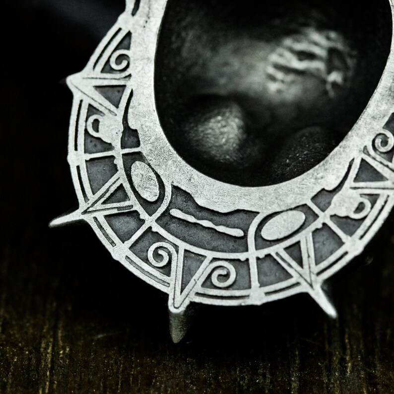 May include: A silver-colored metal pendant with a circular design featuring intricate details and a raised center. The pendant has a sharp, pointed edge.