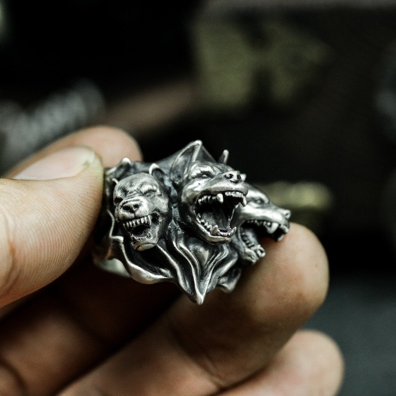 Puede incluir: Un anillo de tono plateado con un dise&ntilde;o detallado de tres cabezas caninas. Cada cabeza est&aacute; esculpida con dientes descubiertos, orejas puntiagudas y detalles de pelaje texturizado. El metal tiene un acabado oxidado que resalta los contornos profundos y los rasgos marcados de las caras de animales gru&ntilde;endo dispuestas alrededor de la banda.