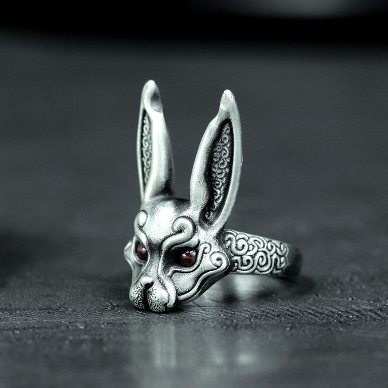 May include: A silver ring with a detailed rabbit head design. The rabbit has long ears and red eyes. The ring band is also decorated with a swirling pattern.