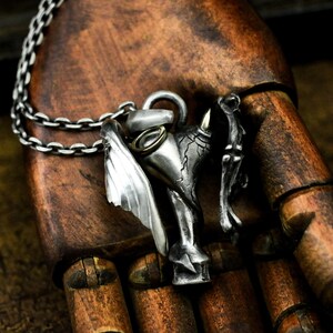 Puede incluir: Un collar con un colgante de plata que presenta una escultura detallada de una criatura alada con cabeza de toro. El colgante est&aacute; unido a una cadena de plata.