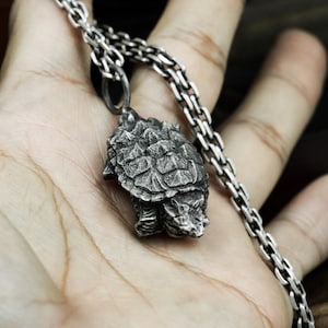 May include: A silver chain necklace with a detailed, three-dimensional pendant of a snapping turtle. The pendant is made of metal and has a realistic texture.
