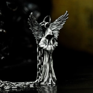 May include: Silver pendant necklace featuring a hooded figure with wings. The figure is standing with its arms crossed and has a detailed, textured design.