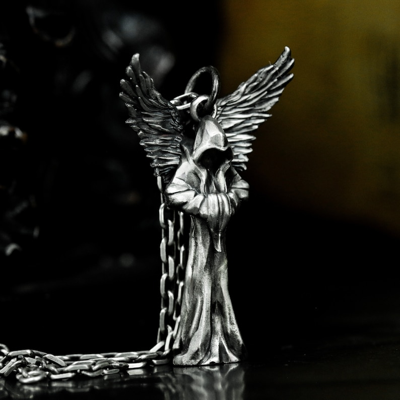 May include: Silver pendant necklace featuring a hooded figure with wings. The figure is standing with its arms crossed and has a detailed, textured design.