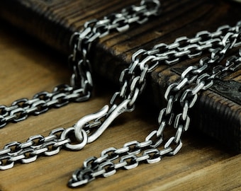 May include: A silver chain necklace with a lobster clasp closure. The chain is made of small, square links. The chain is lying on a wooden surface.