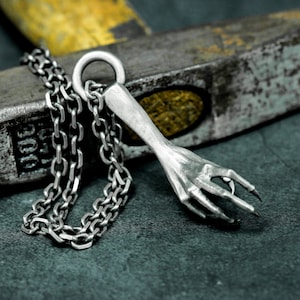 May include: A silver chain necklace with a pendant shaped like a hand with long, sharp claws. The pendant is resting on a gray surface.
