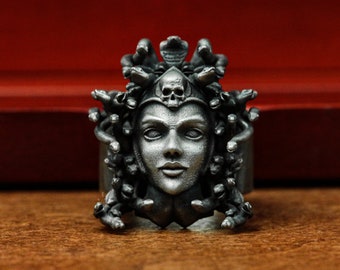May include: A silver ring with a detailed sculpture of Medusa's head. The sculpture features a skull on top of her head and snakes surrounding her face.