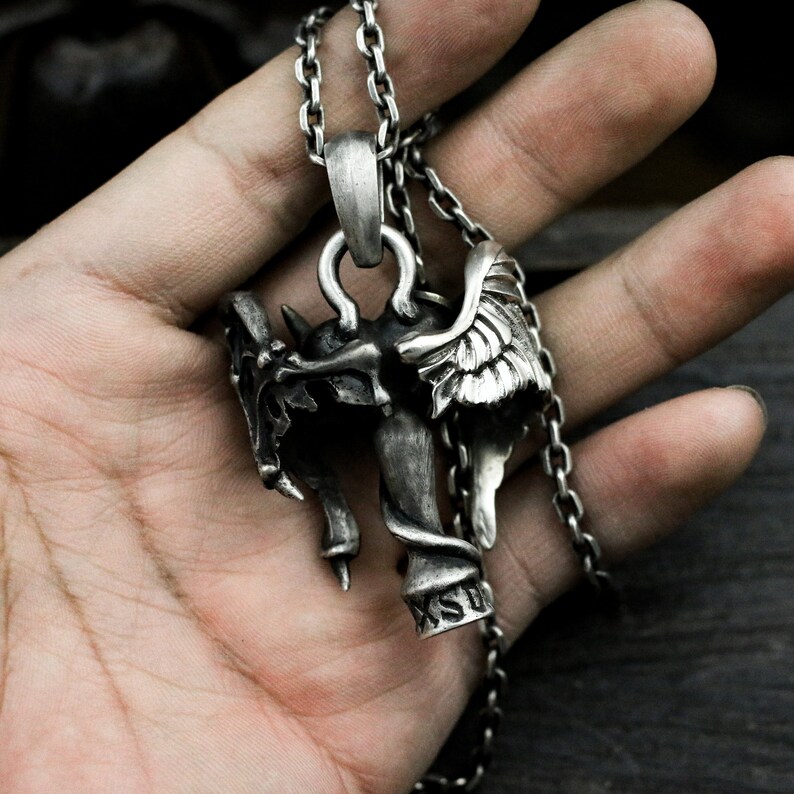Puede incluir: Collar de cadena de plata con un colgante que representa una figura de demonio alado con una cabeza de calavera y una serpiente envuelta alrededor de su cuerpo. El colgante est&aacute; inscrito con las letras "KSD".