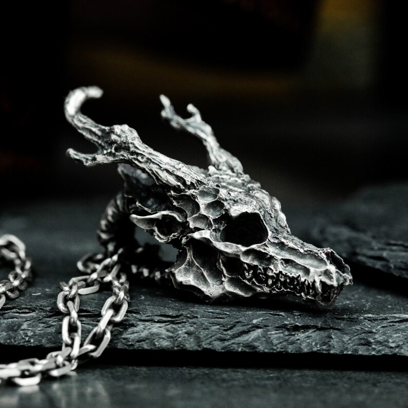 May include: A silver pendant necklace featuring a detailed skull with antlers. The skull is textured and has a realistic look. The necklace is made of a silver chain.
