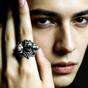 Puede incluir: Un anillo de tono plateado con un dise&ntilde;o de tres cabezas de c&aacute;nido. La banda de metal muestra detalles intrincados en cada una de las tres cabezas gru&ntilde;endo, con un acabado pulido que resalta los rasgos esculpidos de la criatura m&iacute;tica.