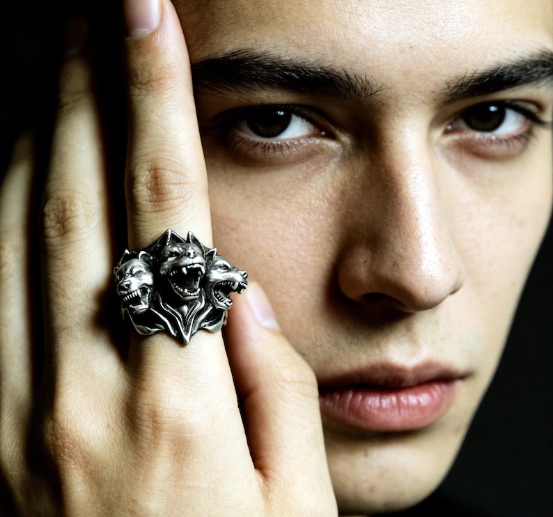 Puede incluir: Un anillo de tono plateado con un dise&ntilde;o de tres cabezas de c&aacute;nido. La banda de metal muestra detalles intrincados en cada una de las tres cabezas gru&ntilde;endo, con un acabado pulido que resalta los rasgos esculpidos de la criatura m&iacute;tica.