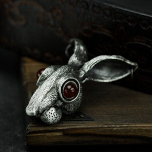May include: A silver rabbit pendant with red gemstone eyes. The pendant is detailed with fur texture and has a long, thin body.