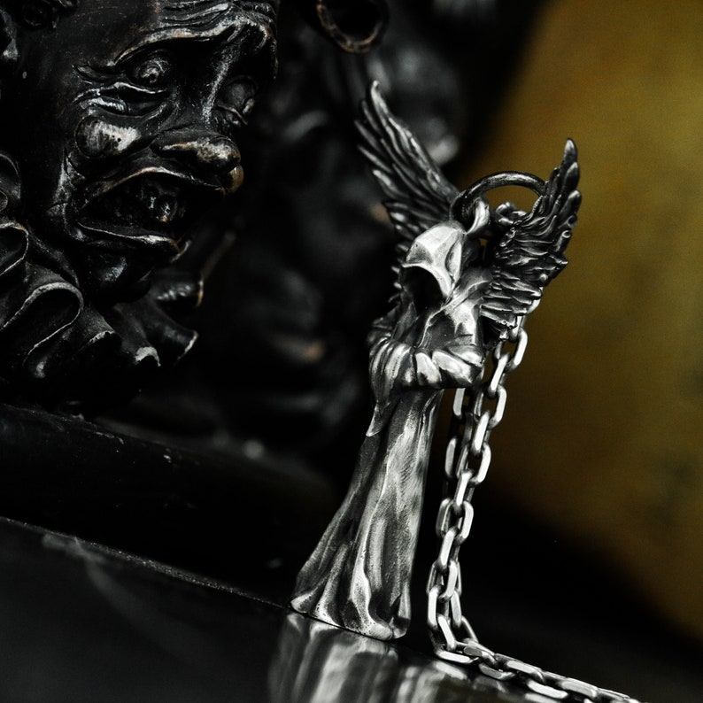 May include: A silver pendant featuring a hooded angel figure with wings, holding a chain in its hands. The angel is standing on a black surface.