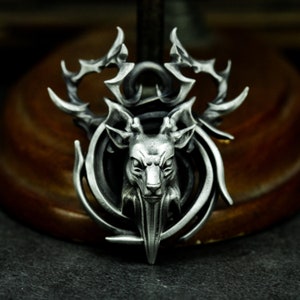 Op de afbeelding: Een zilveren hanger met een gedetailleerd hertenhoofd met geweien, omringd door een wervelend, ingewikkeld ontwerp.