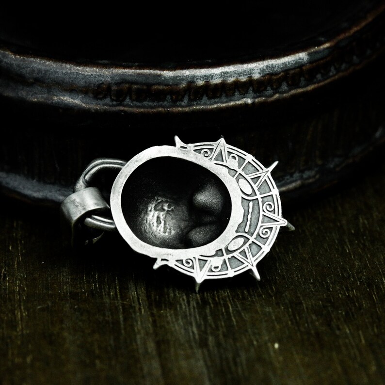 May include: A silver pendant with a detailed design of a face surrounded by a circular pattern with spikes. The pendant has a loop for attaching to a chain.