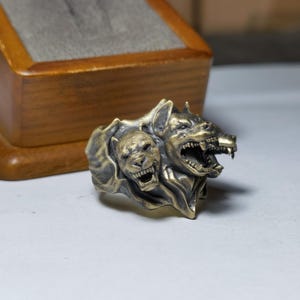 Puede incluir: Un anillo met&aacute;lico con un dise&ntilde;o detallado de tres cabezas caninas con acabado en lat&oacute;n antiguo. La escultura muestra tres cabezas gru&ntilde;endo con dientes visibles y pelaje texturizado, integradas en una sola banda con una p&aacute;tina oscura.