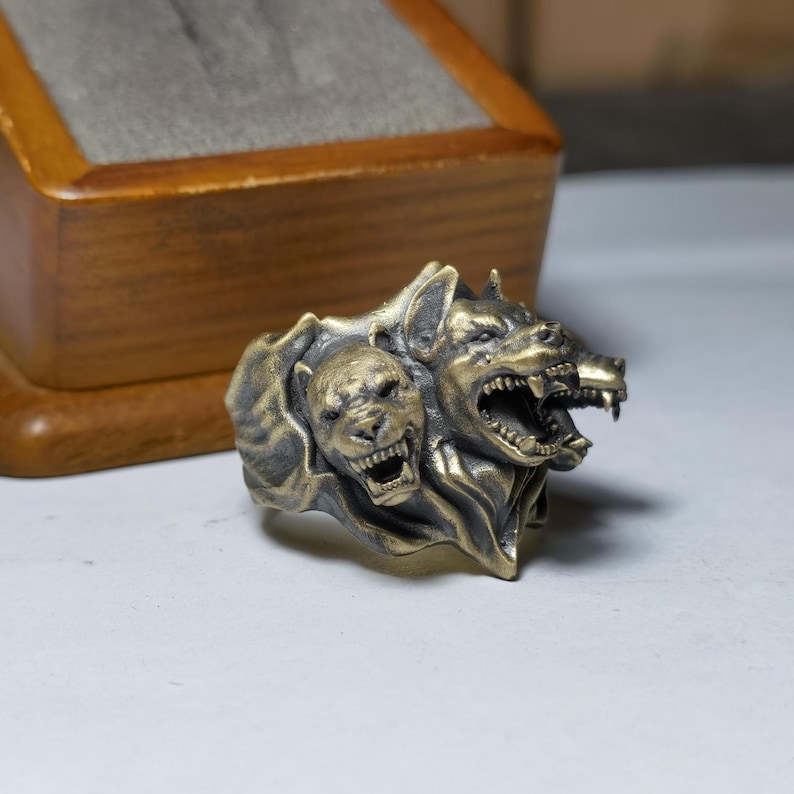 Puede incluir: Un anillo met&aacute;lico con un dise&ntilde;o detallado de tres cabezas caninas con acabado en lat&oacute;n antiguo. La escultura muestra tres cabezas gru&ntilde;endo con dientes visibles y pelaje texturizado, integradas en una sola banda con una p&aacute;tina oscura.