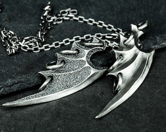 Puede incluir: Un collar de plata con un colgante en forma de ala texturizado y detallado. El colgante está hecho de dos piezas, una con una forma más puntiaguda y la otra con una forma más redondeada. El collar está hecho de una cadena de plata.