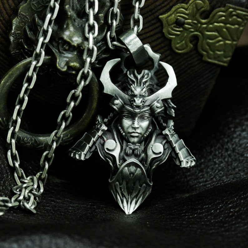 Puede incluir: Un collar con un colgante de plata que representa un busto detallado de un guerrero samur&aacute;i que lleva un casco con un dise&ntilde;o de drag&oacute;n. El colgante est&aacute; en una cadena de plata.
